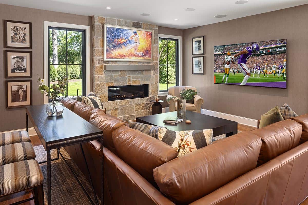 Sports-themed den with leather sectional, stone fireplace, and wall-mounted TVs