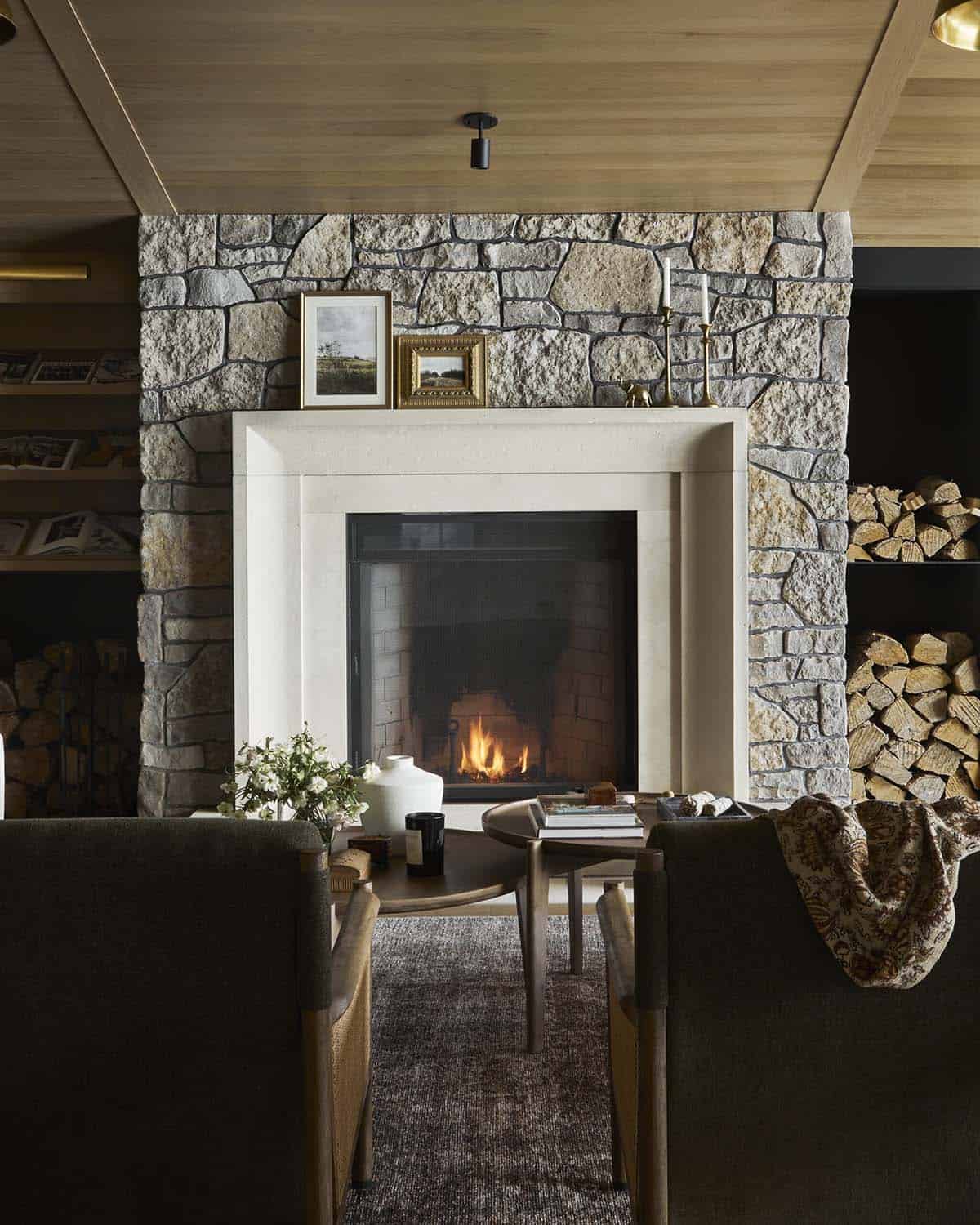 Stone fireplace with lit fire, flanked by log storage, framed by lounge chairs
