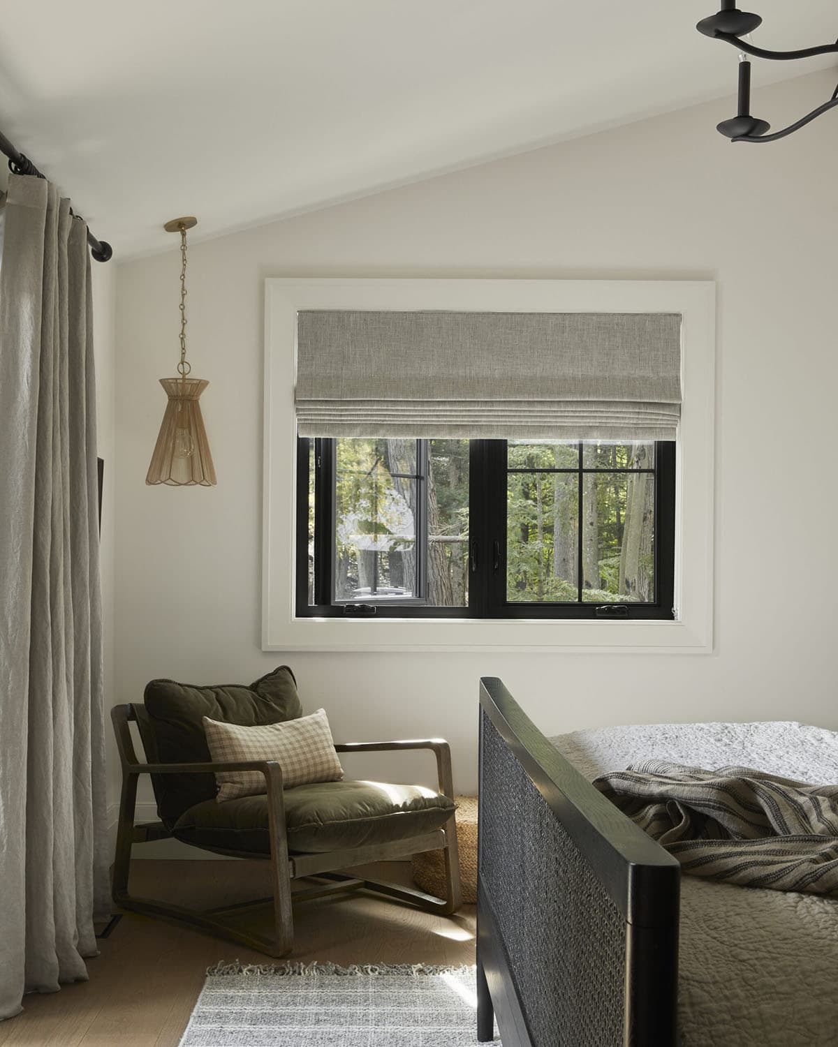 Bedroom corner with olive lounge chair, black cane bed frame, and forest-view window