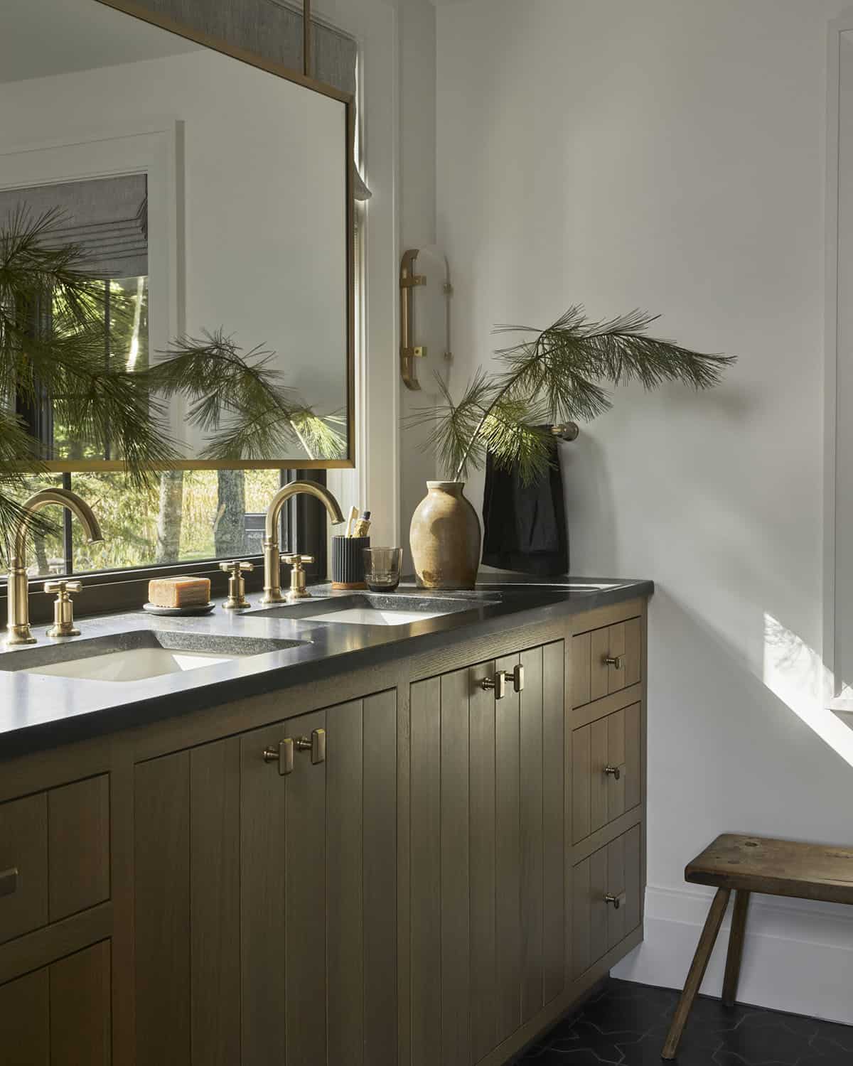 Bathroom vanity with brass fixtures, pine branch in ceramic vase, and wood stool