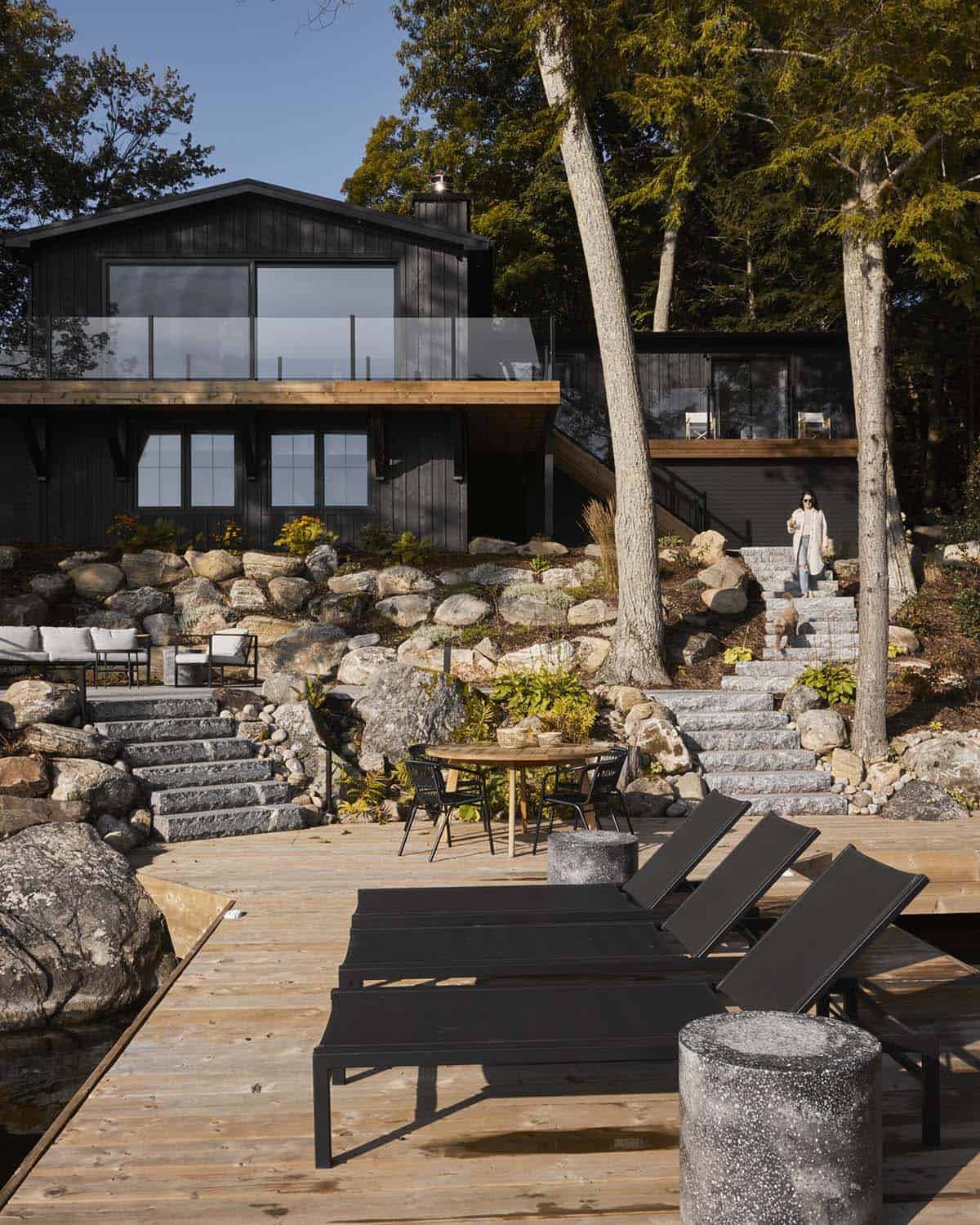 Exterior view of black board-and-batten cottage with stone steps down to lakeside dock and lounge chairs