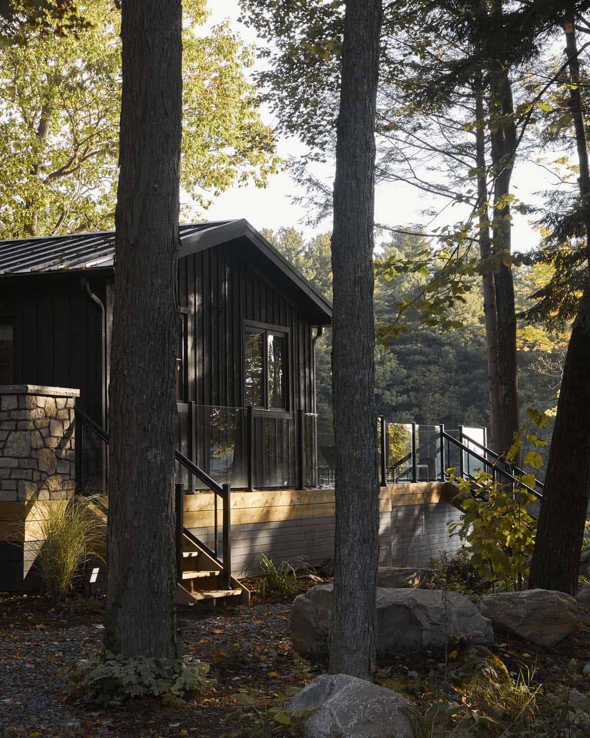Black cottage exterior seen through forest trees with wood deck and glass railings