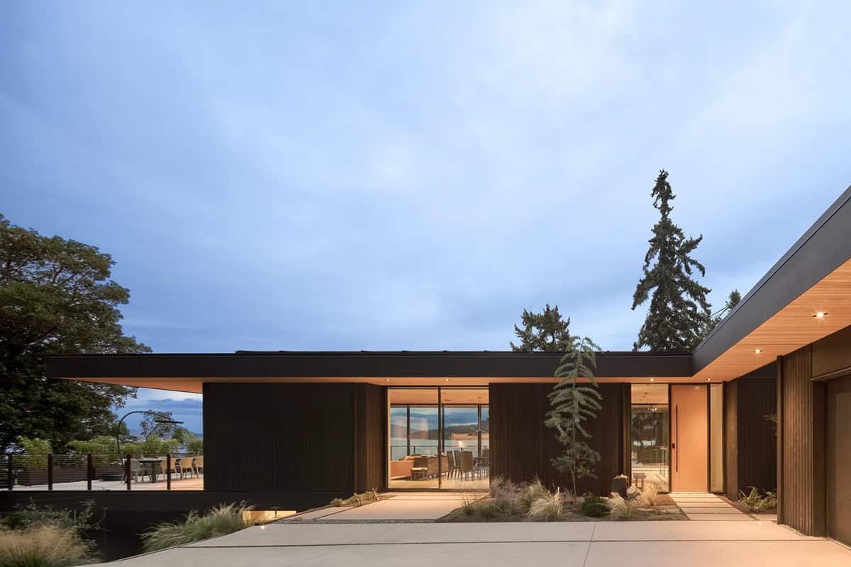 Layer House entry courtyard at dusk with glowing wood soffit and glimpse of inlet beyond