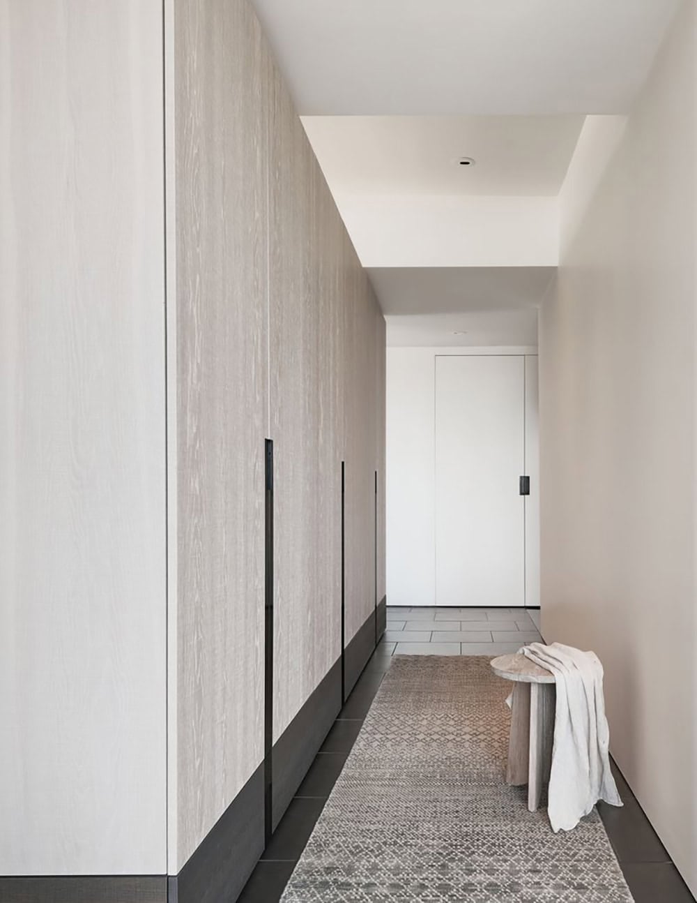 Minimalist hallway with floor-to-ceiling white oak cabinetry, black pull handles, dark tile floor, and a patterned runner rug leading to a white entry door