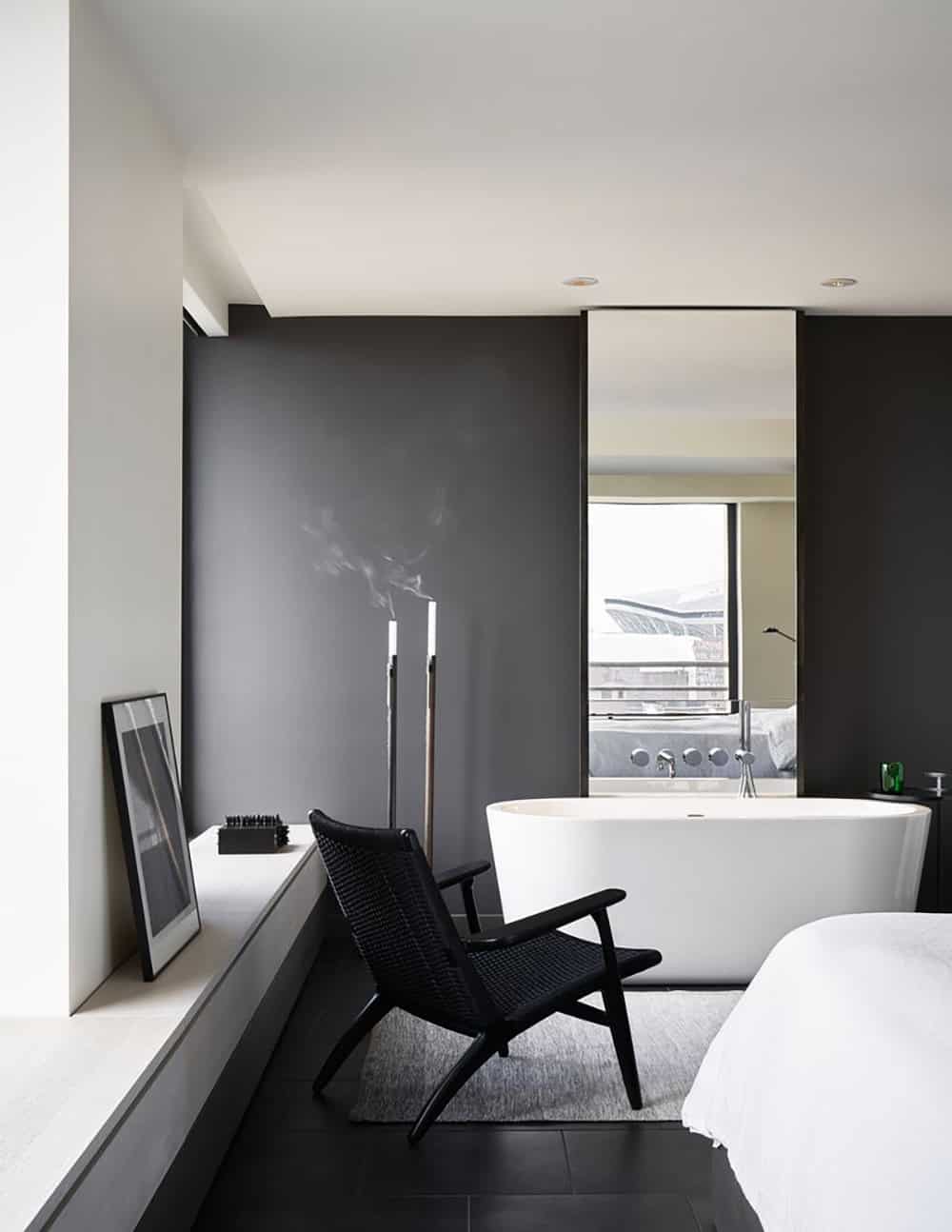 Moody primary bedroom corner with dark gray walls, freestanding white soaking tub, black lounge chair, and tall candlesticks against a large mirror