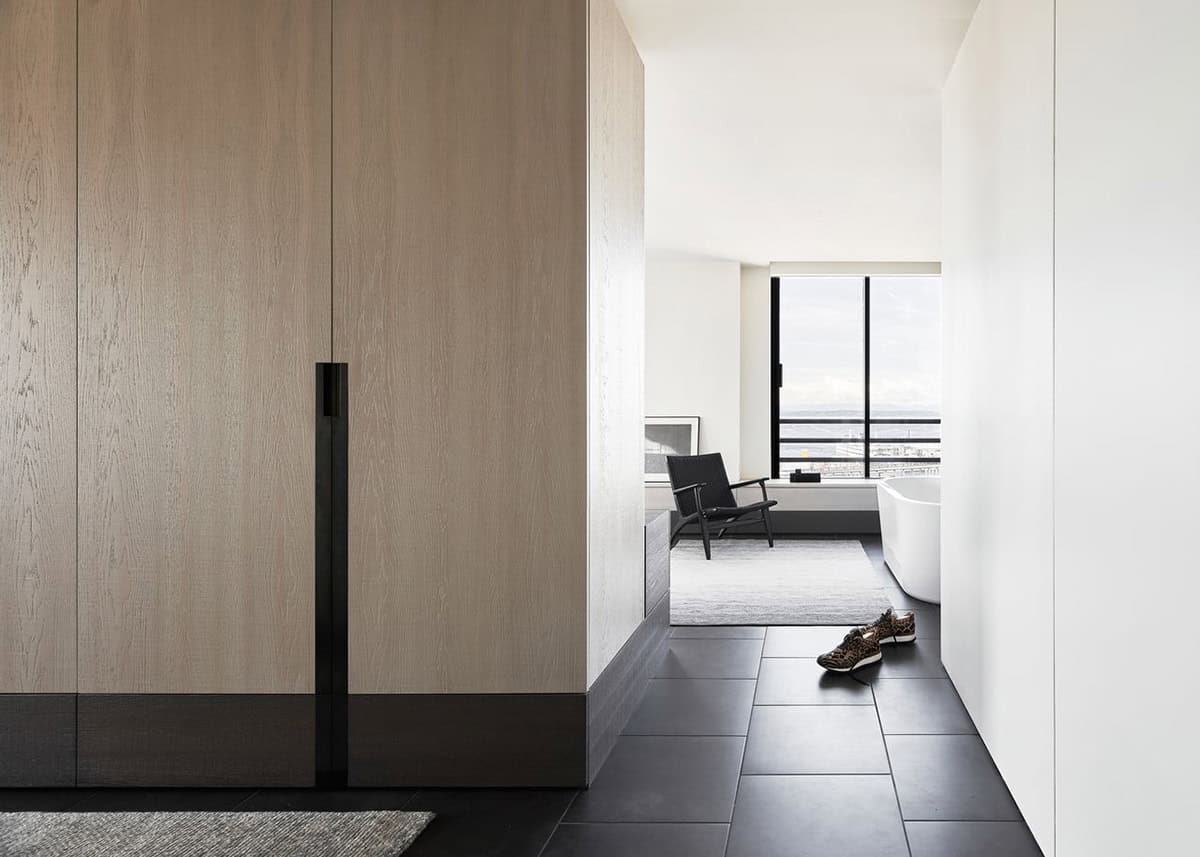 Close-up of white oak wardrobe panels with slim black handles opening to a bedroom with city views and a freestanding tub