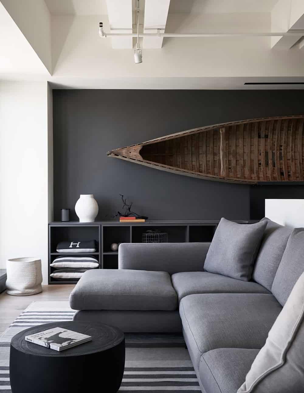 Living room with large gray sectional sofa, black round coffee table, striped rug, dark shelving unit, and an antique wooden canoe mounted on a charcoal accent wall