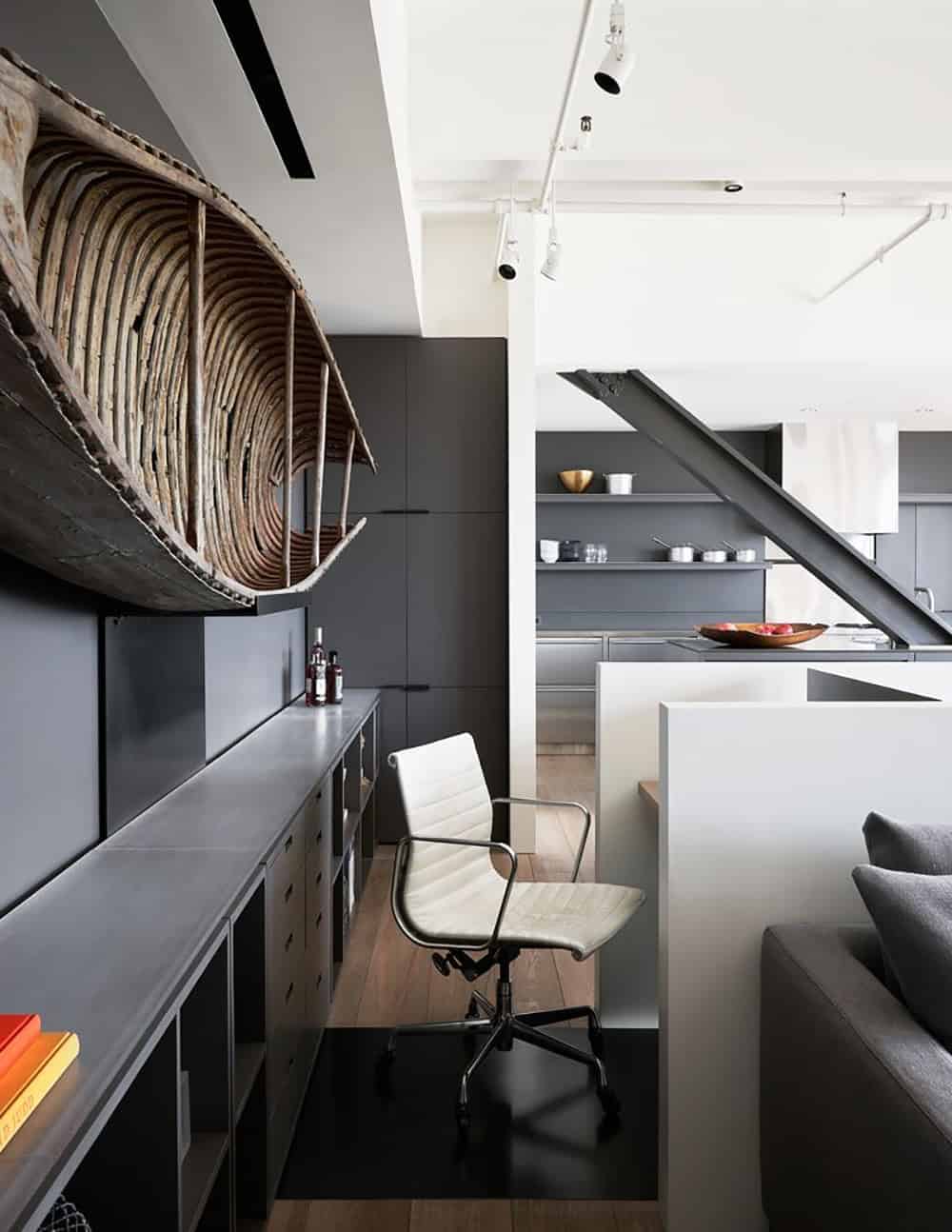Open-plan interior showing the mounted canoe above built-in shelving, a white office chair at a steel-topped desk, and a dark kitchen beyond a structural steel staircase beam