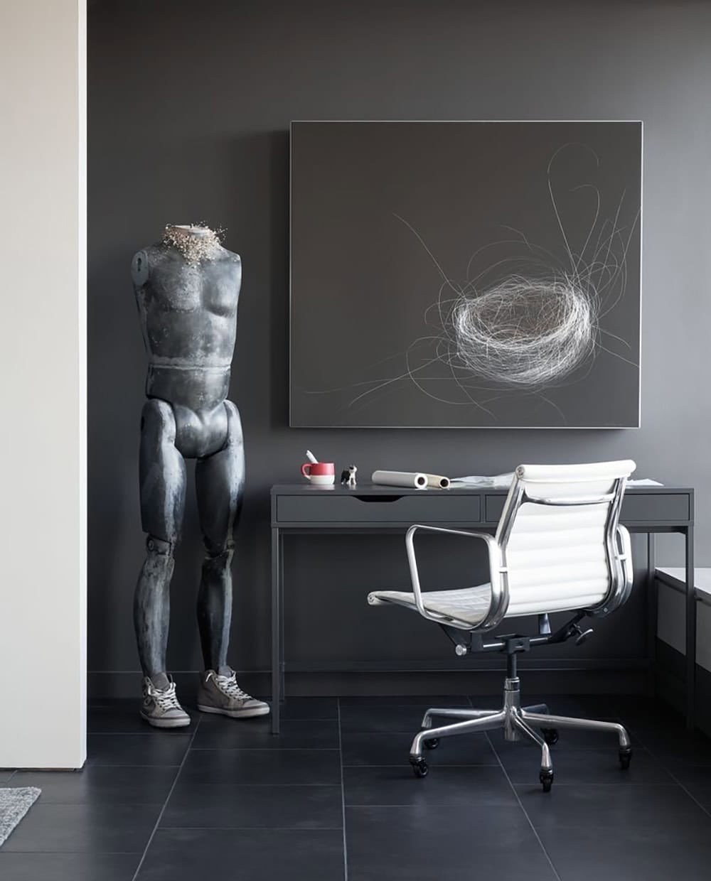 Home office nook with charcoal walls, a gray metal desk, white Eames-style office chair, abstract wire artwork, and a weathered mannequin torso sculpture
