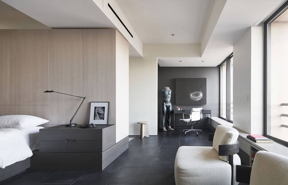 Bedroom suite overview showing light oak headboard wall, dark nightstand, sculptural mannequin in the study nook beyond, cream armchair by floor-to-ceiling windows