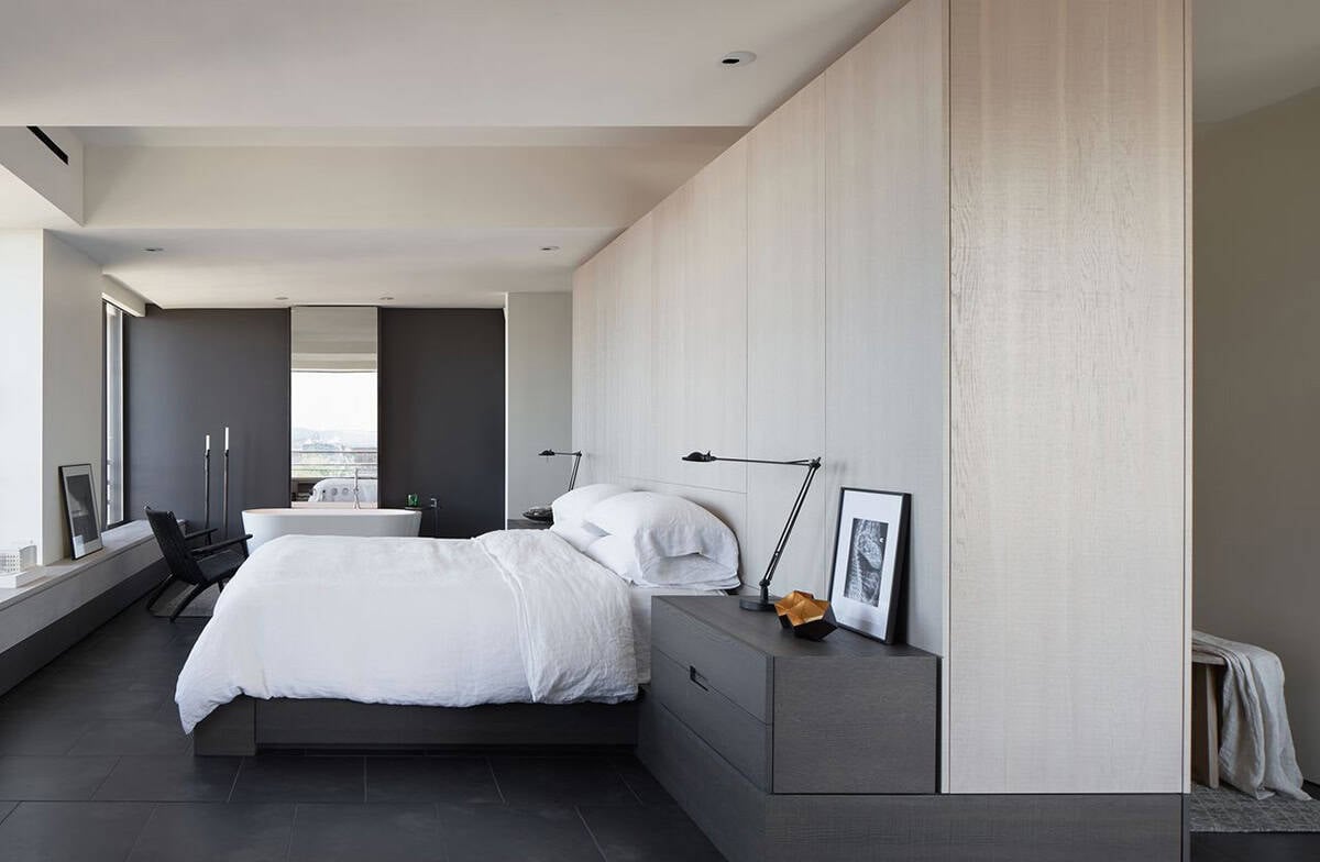 Primary bedroom with white linen bedding, dual Tizio desk lamps, tall white oak wardrobe wall, and open view to the freestanding tub and dark accent wall beyond