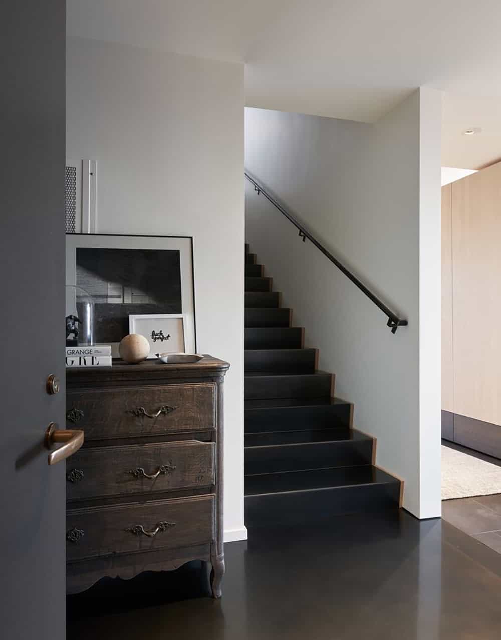 Staircase foyer with dark-painted steps, slim black metal handrail, an ornate antique French commode, and layered black-and-white framed artwork