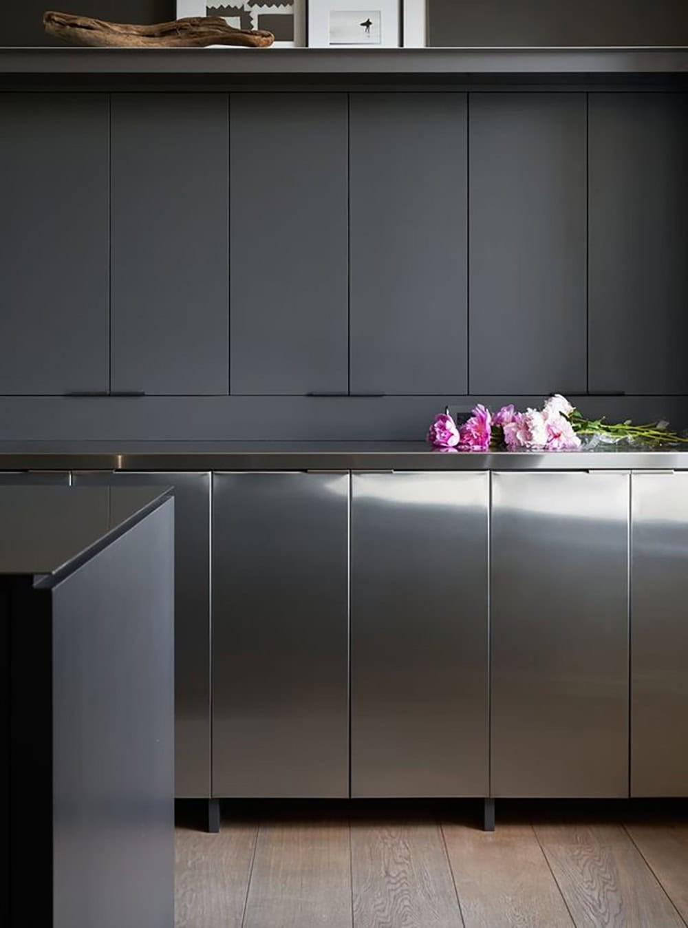 Dark kitchen detail with matte charcoal upper cabinets, brushed stainless steel lower cabinets and countertop, and fresh pink peonies on the counter