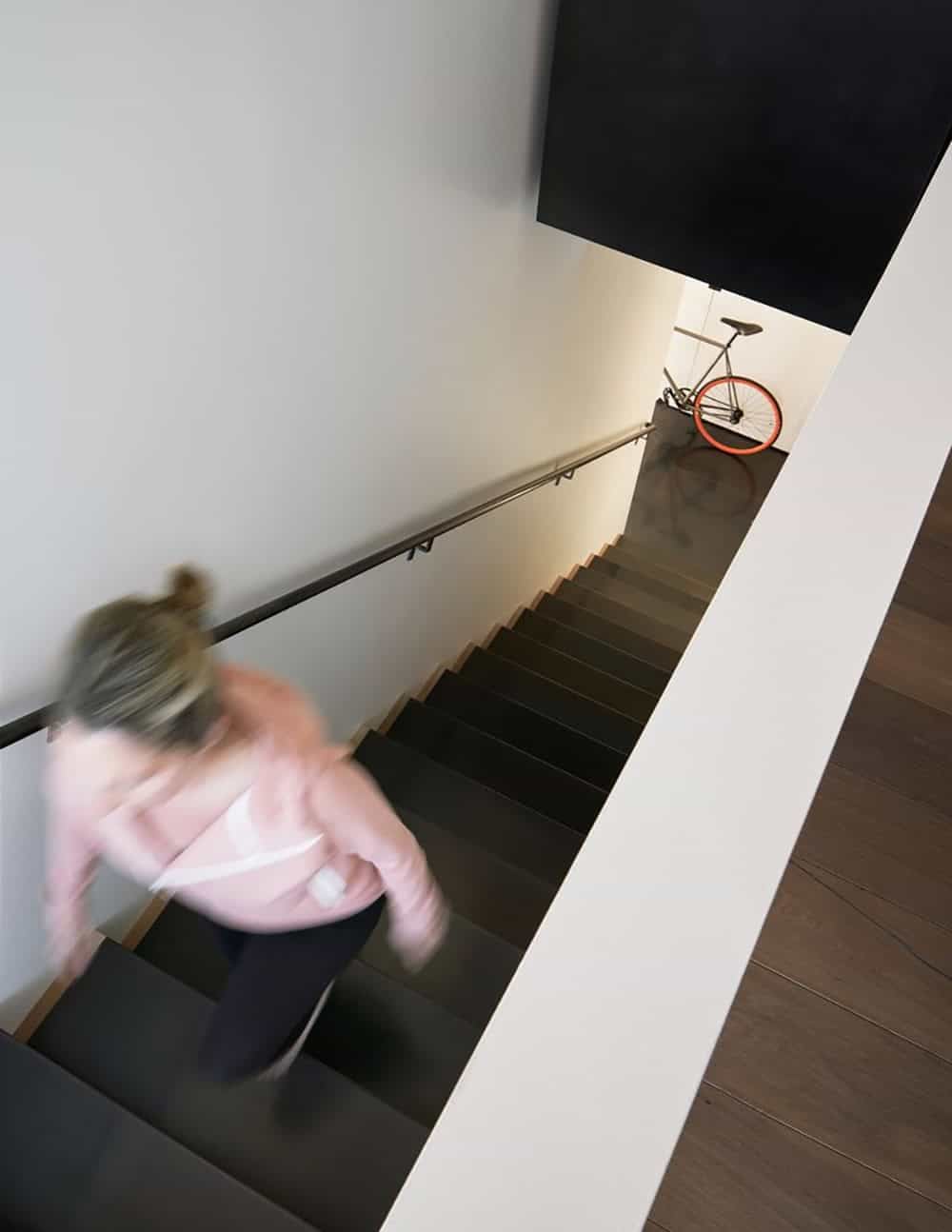 Aerial view looking down a dark staircase with a slim metal handrail, a blurred figure ascending, and an orange-wheeled bicycle leaning at the bottom