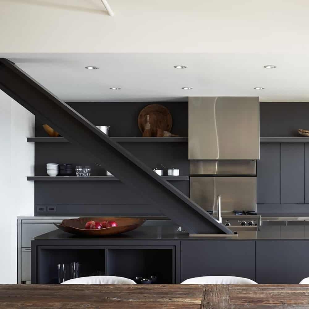 Dark kitchen with matte gray cabinetry, open shelving, stainless steel range hood, and a bold structural steel stair beam cutting diagonally across the space