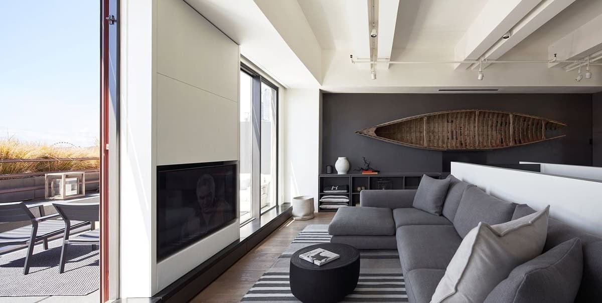 Open-plan living room with floor-to-ceiling windows overlooking a rooftop terrace, a gray sectional sofa, wall-mounted flatscreen TV, and a canoe on the dark accent wall