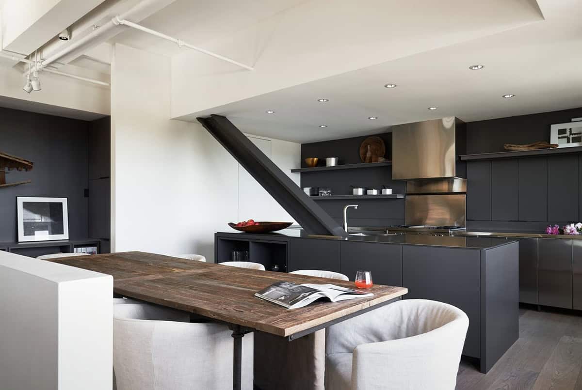 Kitchen and dining area with dark cabinetry, stainless steel countertops, reclaimed wood dining table, cream barrel chairs, and a diagonal steel stair structure