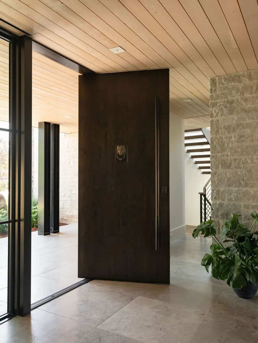 Dark wood pivot front door with brass lion knocker, flanked by stone column and wood-clad ceiling