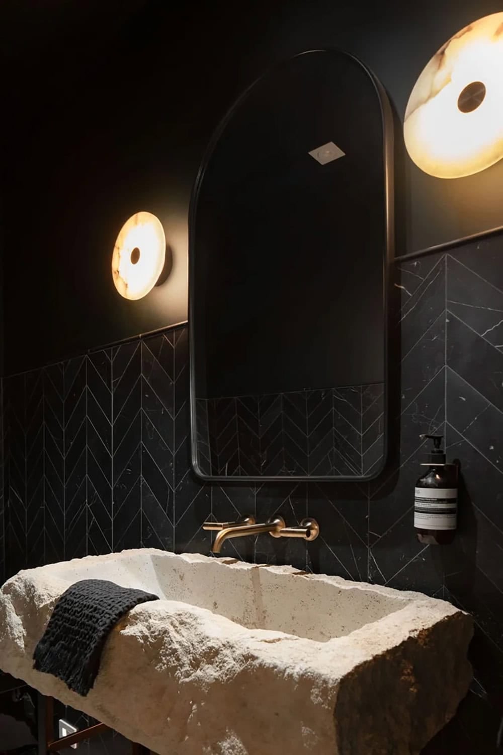 Moody powder room with black herringbone tile, arched black mirror, alabaster sconces, and a rustic stone trough sink