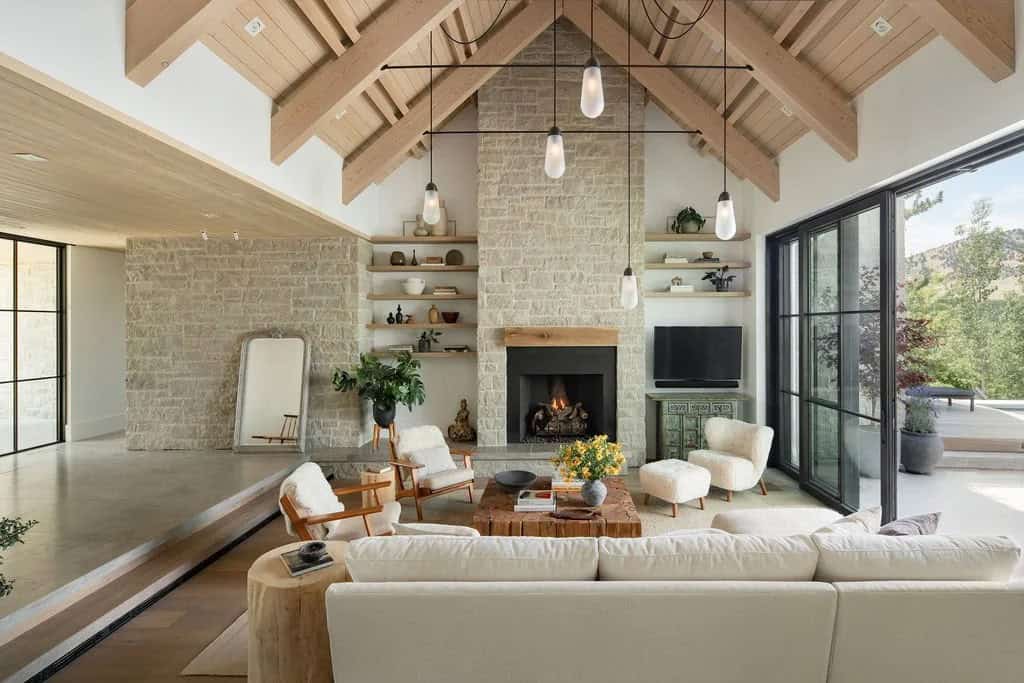 Vaulted living room with exposed wood beams, stone fireplace, open shelving, and cream upholstered chairs