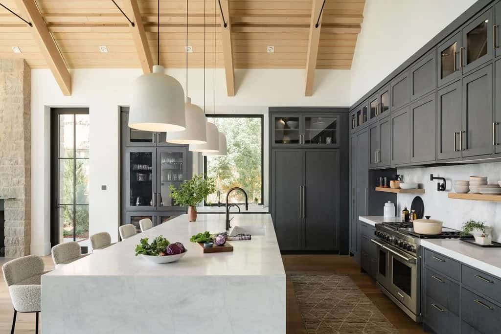 Kitchen with dark gray cabinets, marble island, and wood-beamed ceiling with white pendant lights