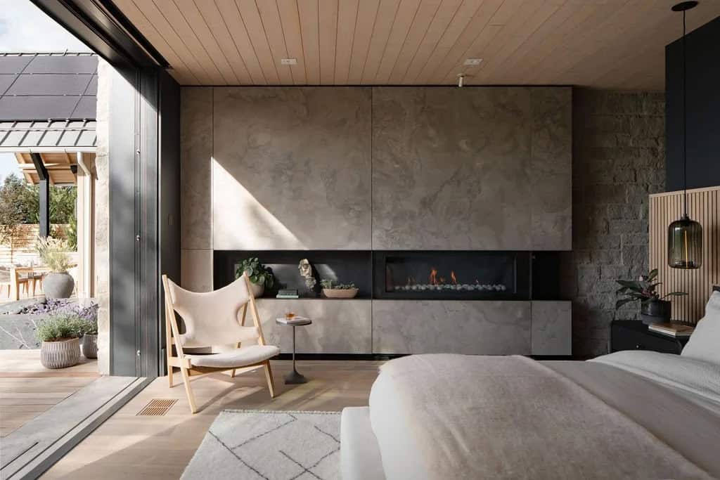 Bedroom with linear gas fireplace in stone-textured wall, leather lounge chair, and view to outdoor patio