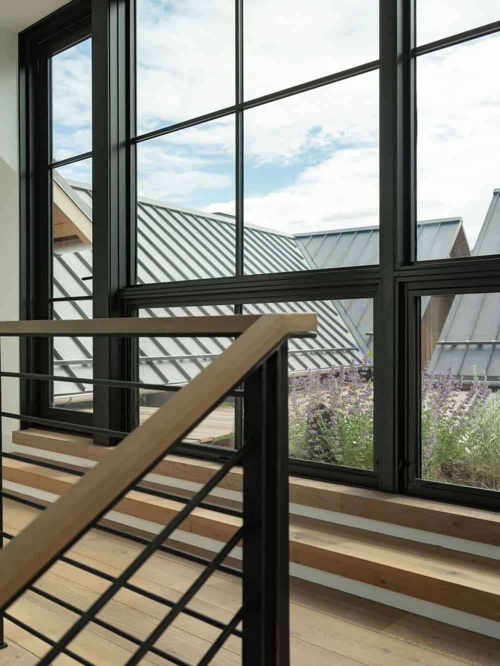 Interior stairwell with wood and steel railing beside floor-to-ceiling windows overlooking rooftops and lavender