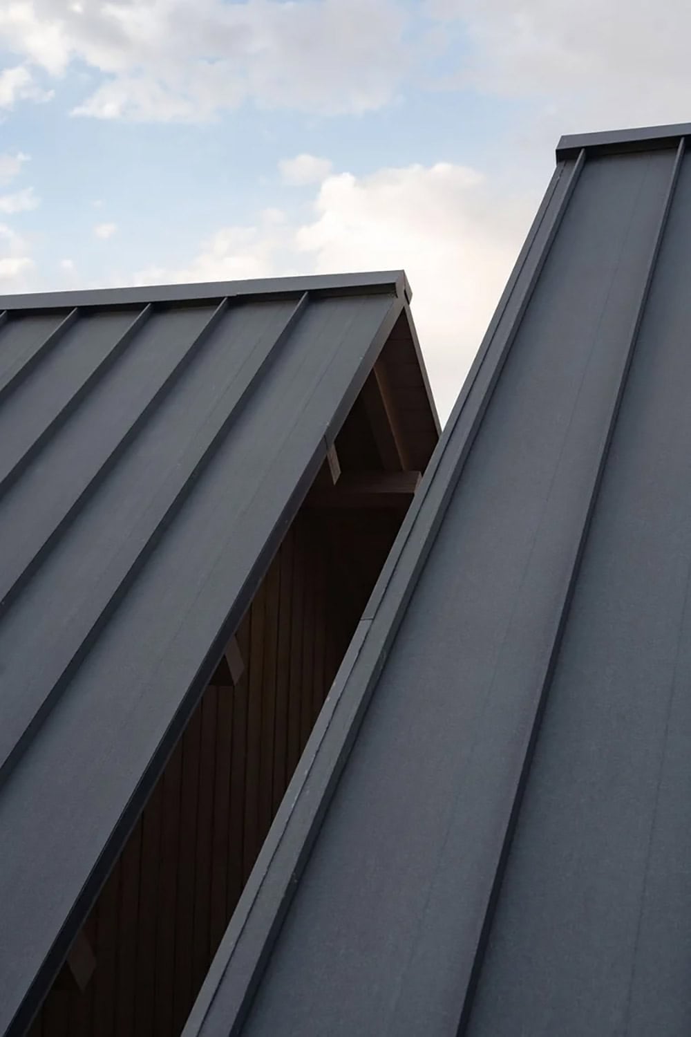 Upward view of dark gray standing-seam metal roof planes meeting against a cloudy sky