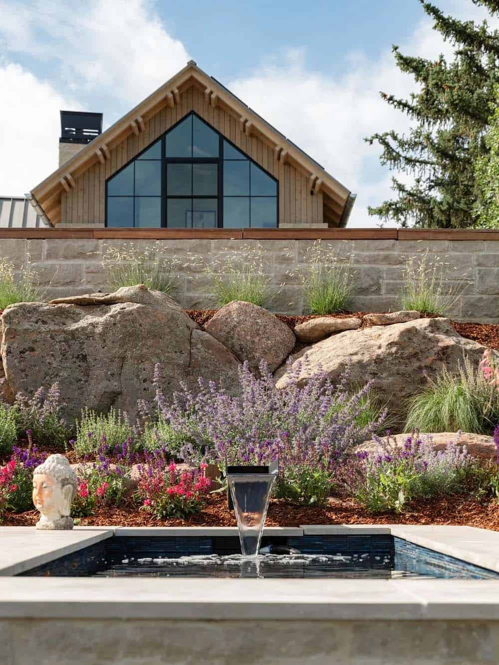Backyard water feature with stone spillway, boulders, and lavender below a gabled glass facade