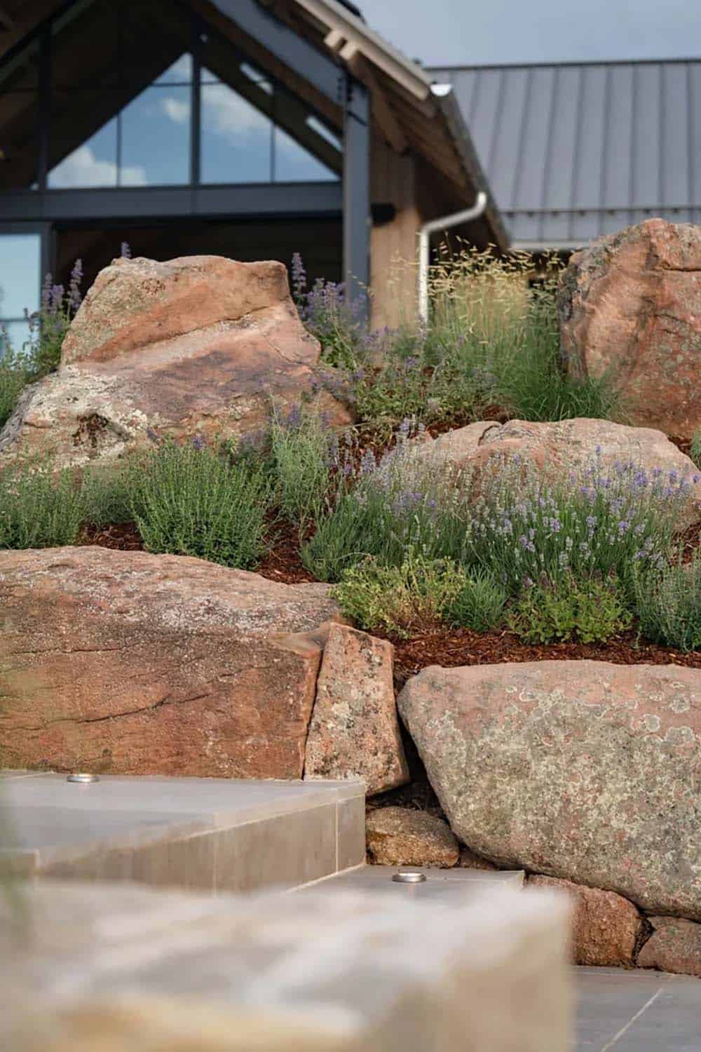Large boulders and lavender plantings beside a stone-edged patio near the home's glass gable