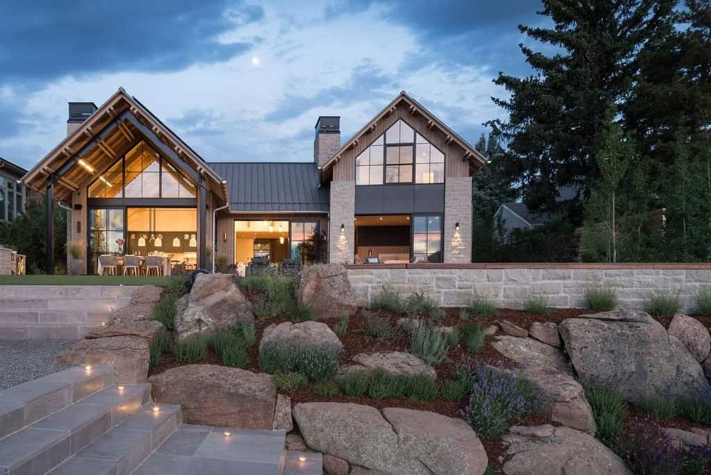 Twilight view of the home's rear facade with illuminated interiors, stone retaining wall, and boulder landscaping