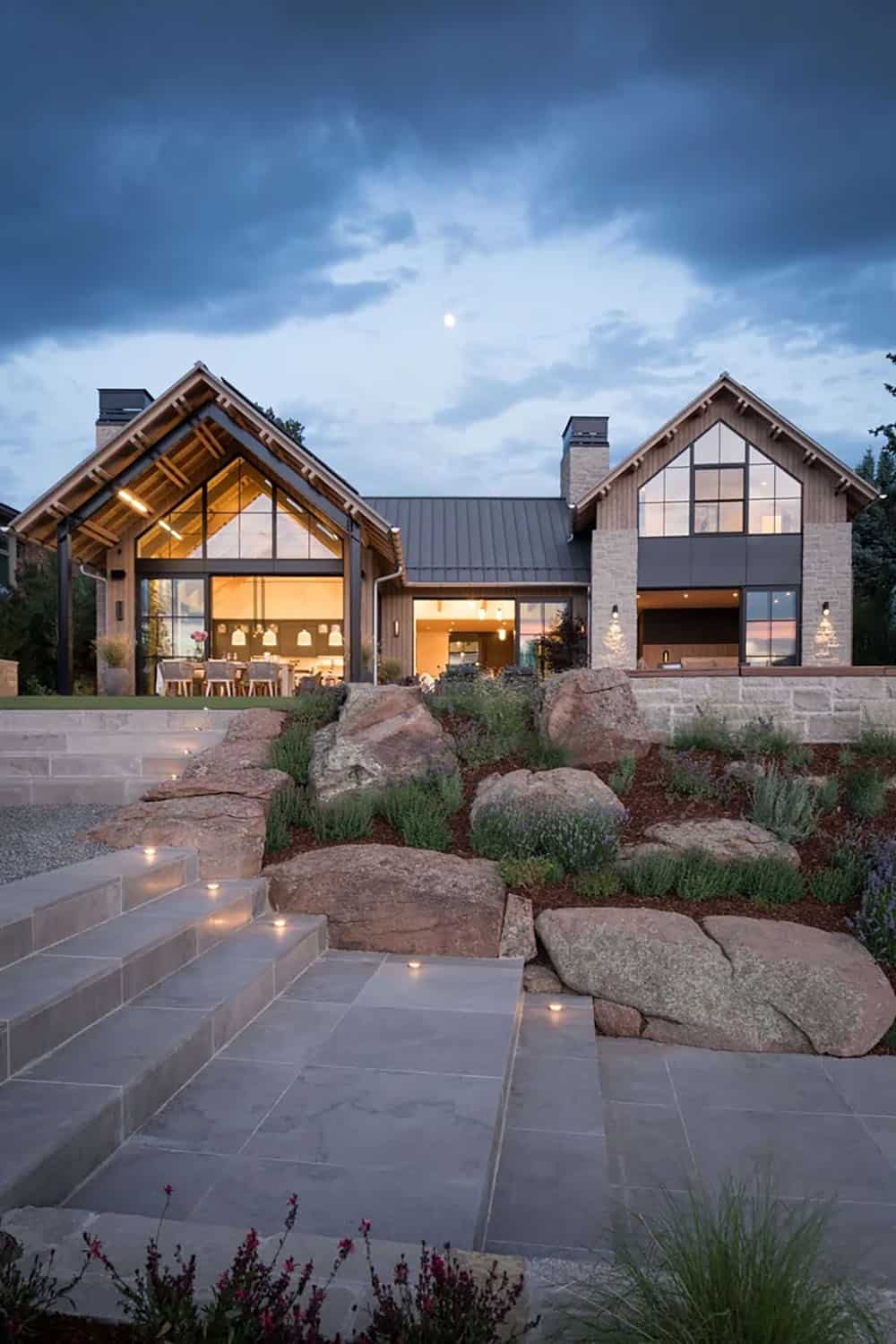 Evening exterior of gabled glass living spaces glowing warmly above a boulder and lavender hillside
