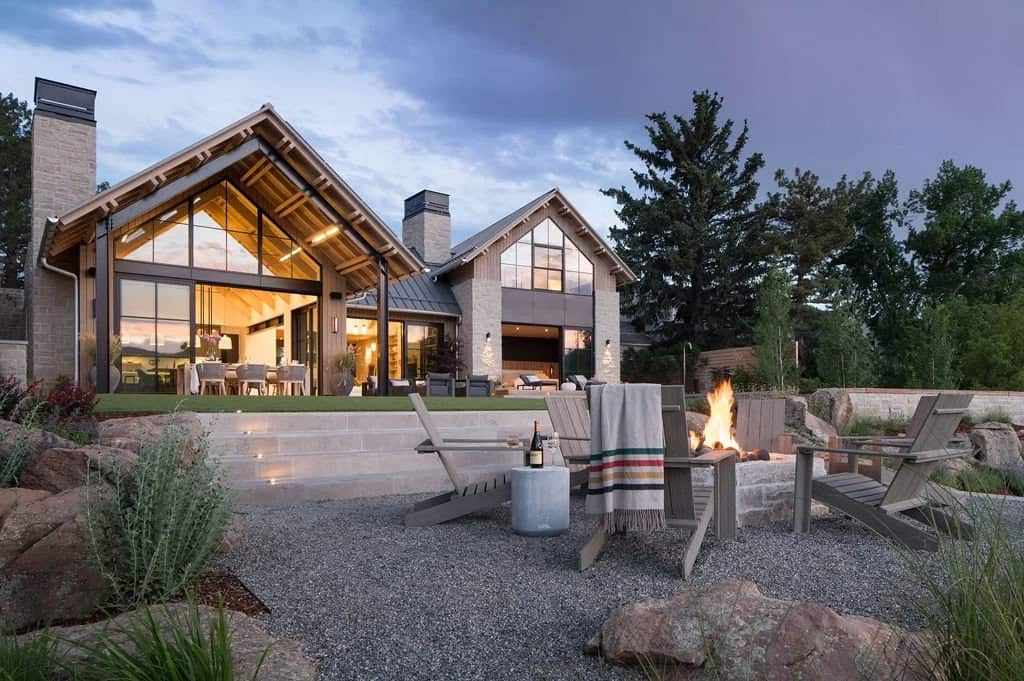 Backyard fire pit and Adirondack chairs on gravel patio facing the illuminated home at dusk