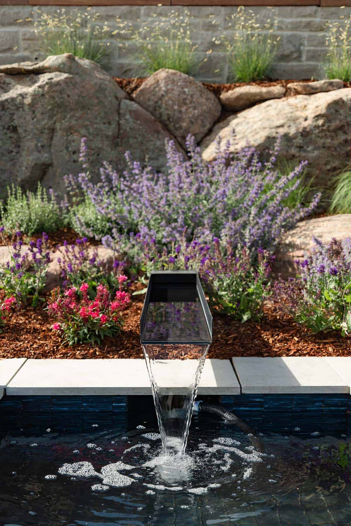 Detail of a sleek black spout pouring water into a glass-tiled pool surrounded by purple flowering plants
