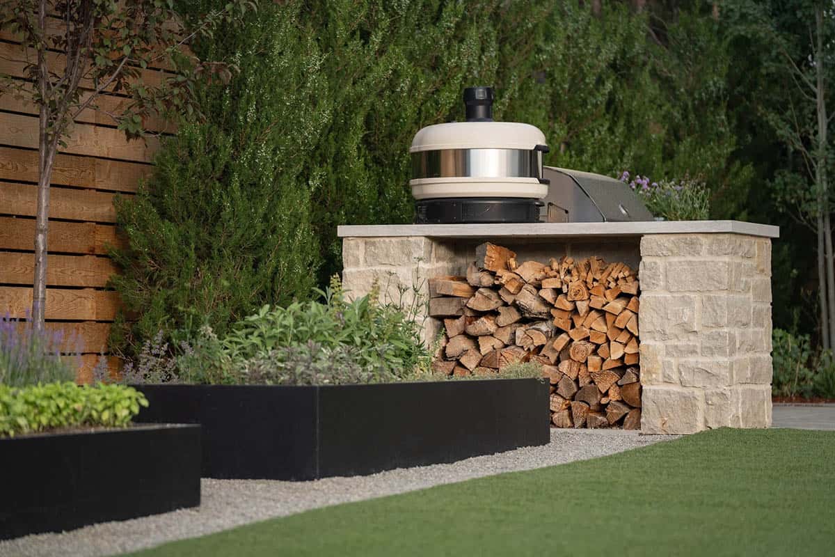 Outdoor kitchen nook with built-in grill, stacked firewood, and stone counter against a cedar fence