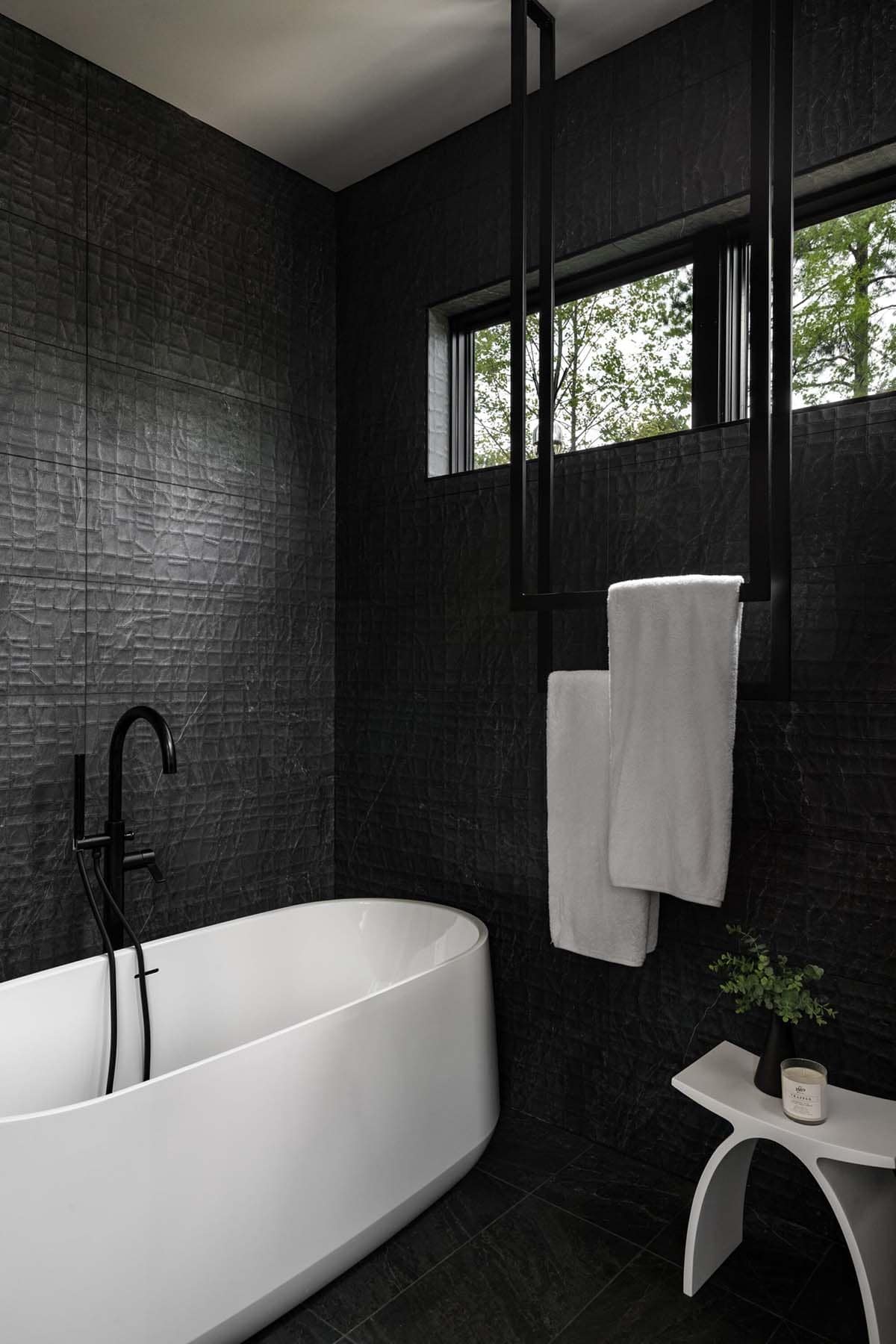 Black-tiled bathroom with freestanding tub and hanging towel bar near window