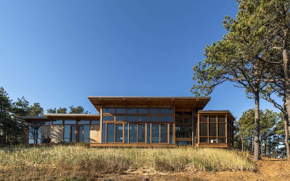 Mid-century inspired beach house with cedar siding and floor-to-ceiling wood-framed windows set among coastal pine trees and dune grass, Massachusetts