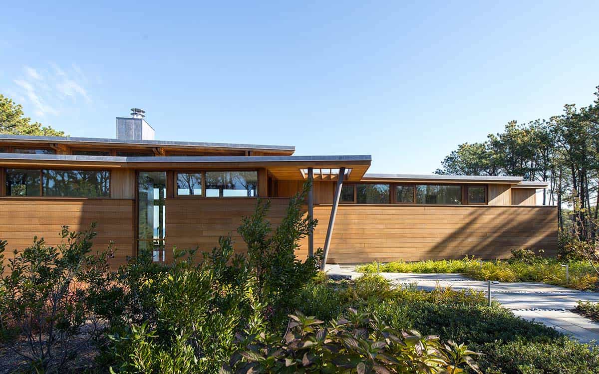 Close-up of the cedar-sided exterior of a mid-century modern beach house with clerestory windows, angled roof canopy, and lush native shrubs