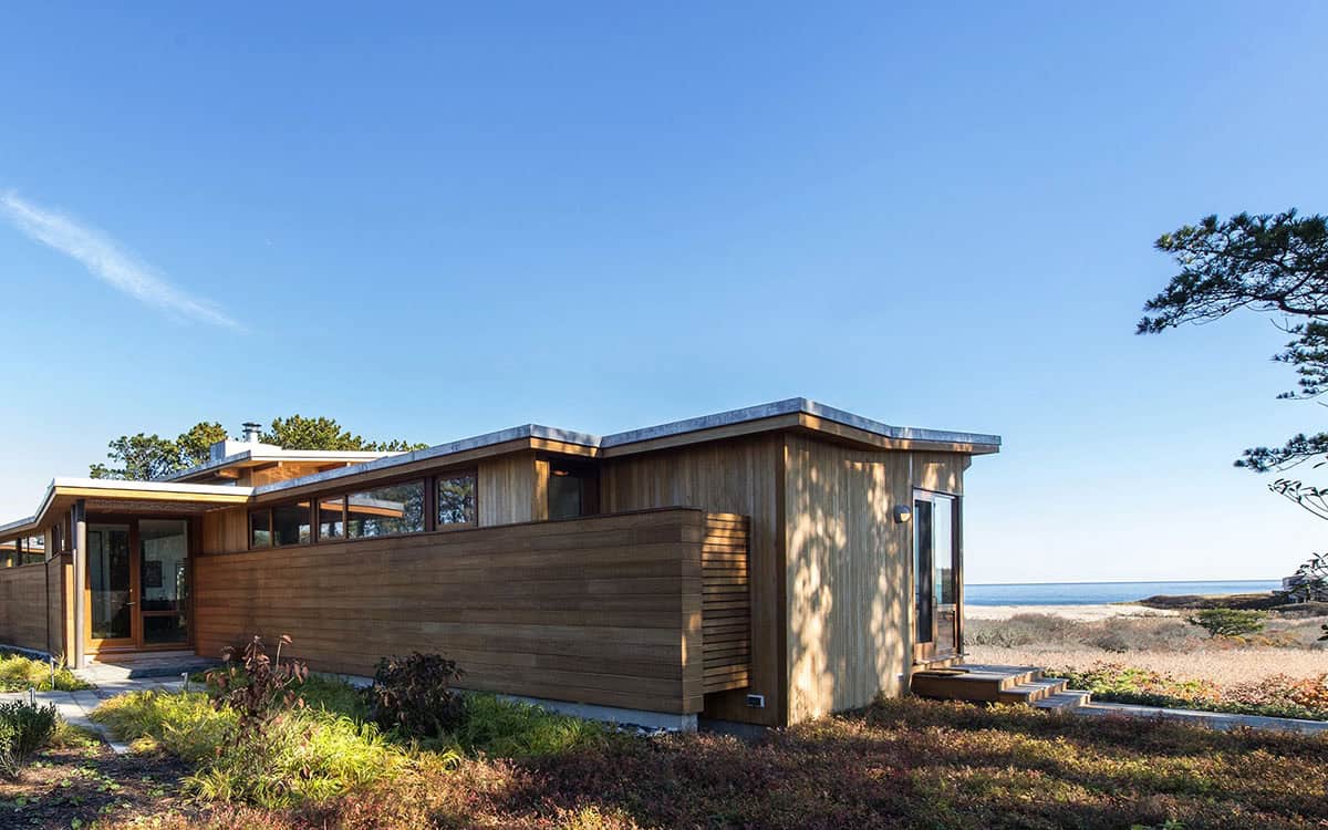 Side view of a cedar-clad mid-century beach house with flat and shed rooflines, vertical siding accent, and ocean visible beyond the dunes