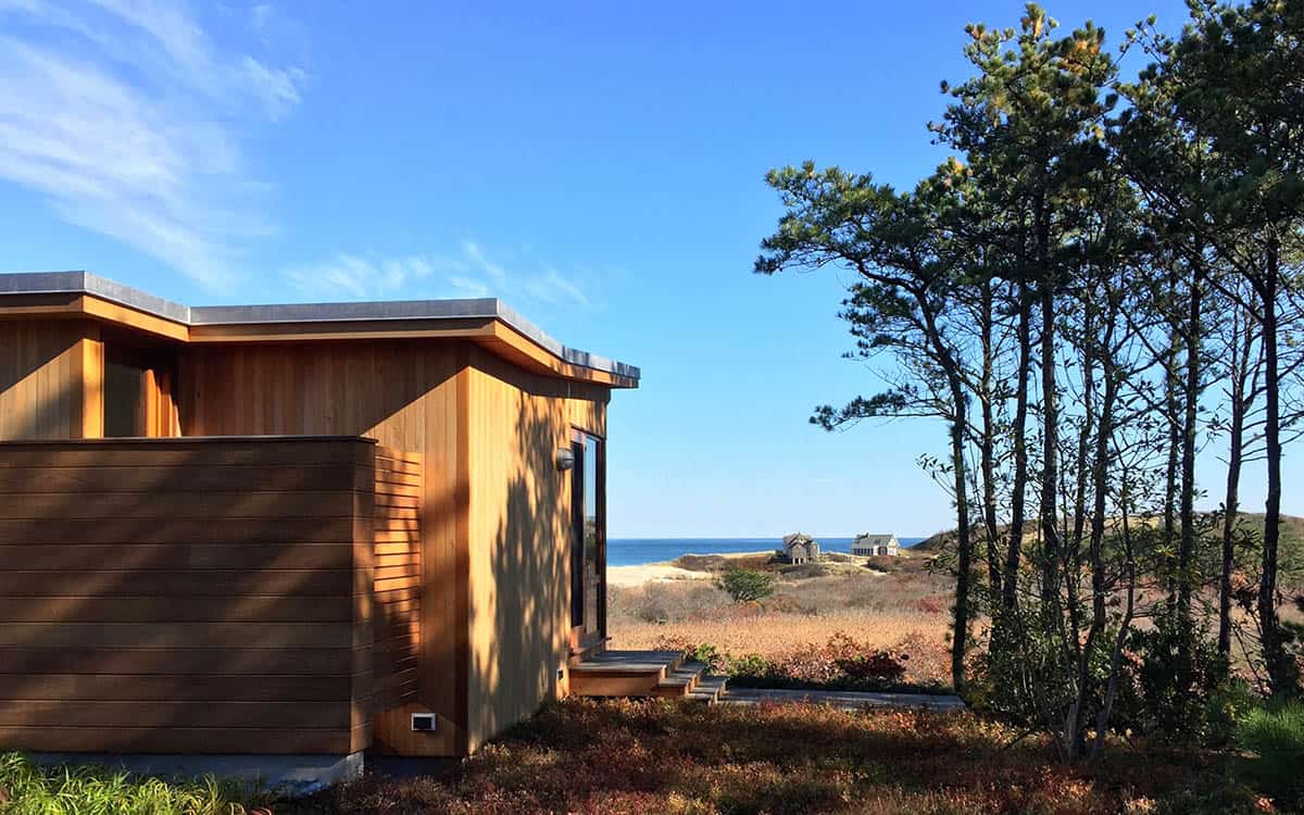 Corner detail of a cedar-sided beach house with flat roof and ocean view beyond dune grass and pitch pine trees at golden hour
