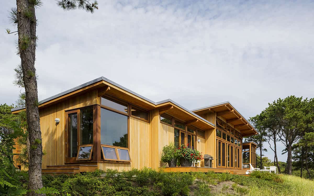 Sunny exterior view of a mid-century modern beach house with vertical cedar siding, large corner windows, and a wood-framed screened porch