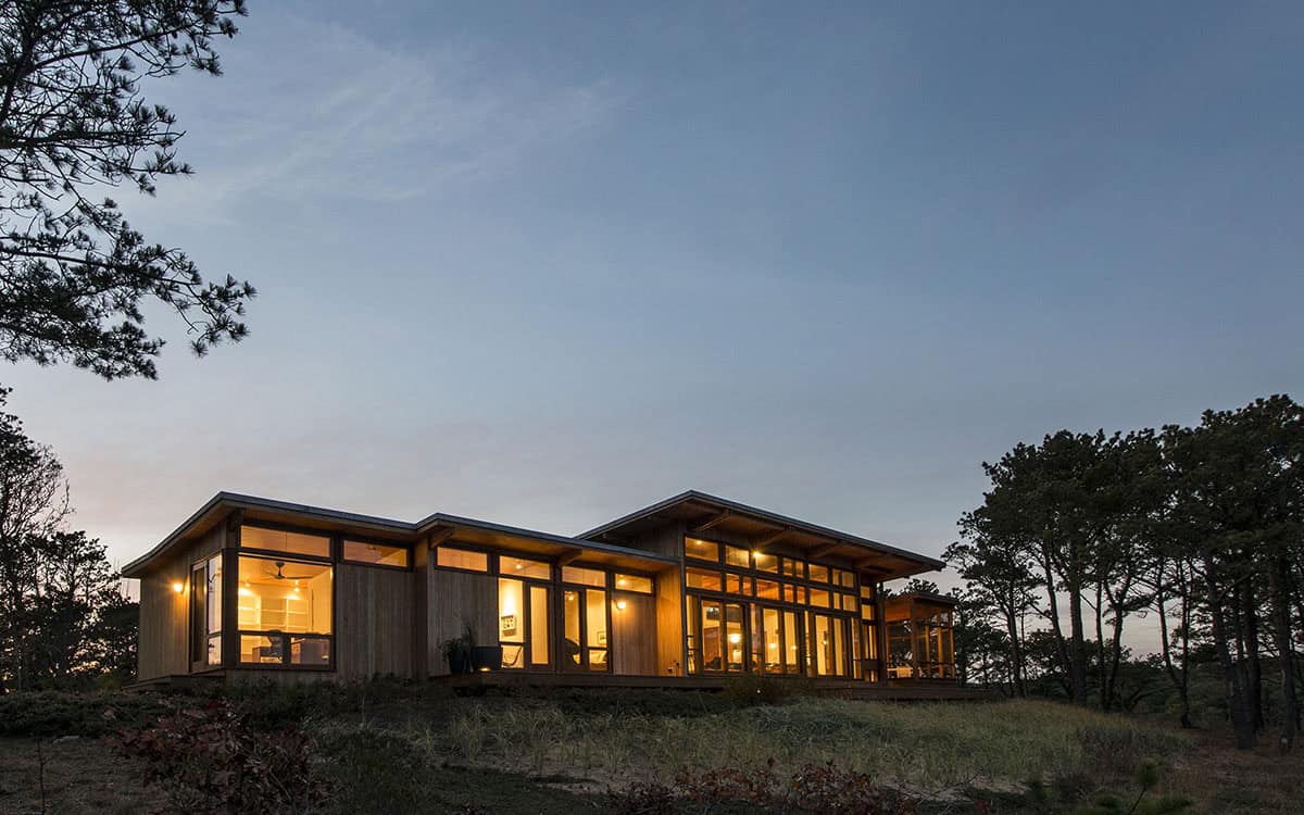 Dusk exterior of a mid-century modern beach house with warm interior light glowing through floor-to-ceiling wood-framed windows, set in a coastal pine landscape