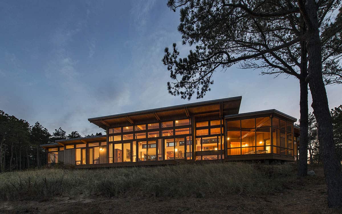Evening exterior of a mid-century inspired home with expansive wood-framed glazing illuminated from within, surrounded by pitch pines and dune grass