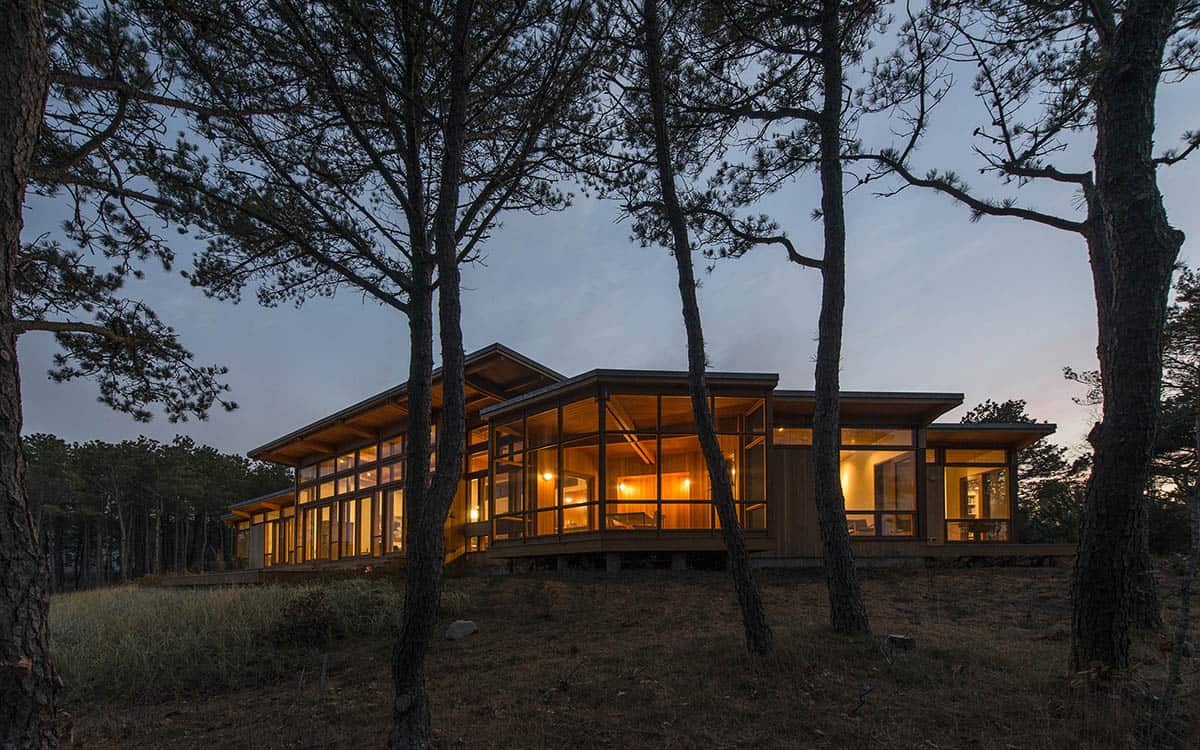 Twilight view of a mid-century modern beach house glowing through floor-to-ceiling windows, framed by tall coastal pines