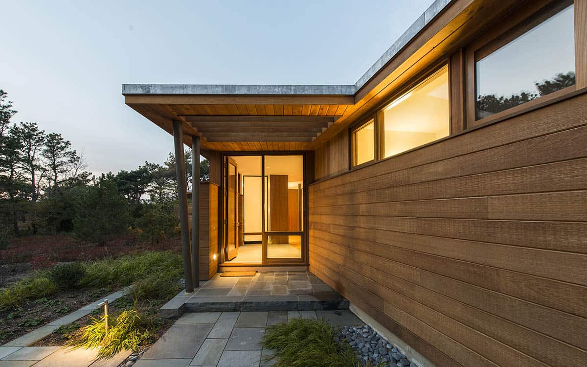 Covered entry of a mid-century modern beach house at dusk, featuring a flat concrete roof overhang, wood-clad ceiling, glass door, and bluestone path
