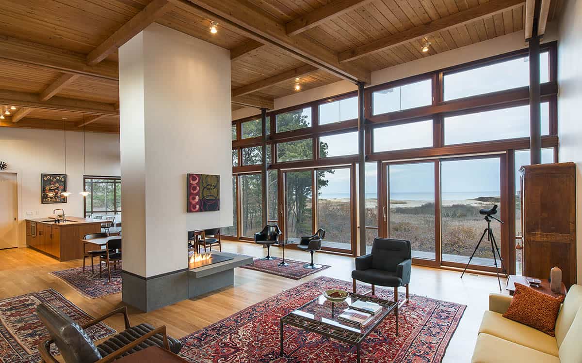 Open-plan living room with exposed wood beam ceiling, double-sided fireplace, black leather chairs, Persian rugs, and floor-to-ceiling windows overlooking coastal dunes and ocean