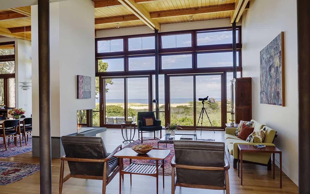 Mid-century modern living room with wood-framed floor-to-ceiling windows, leather lounge chairs, a green sofa, Persian rug, and ocean views through the dunes