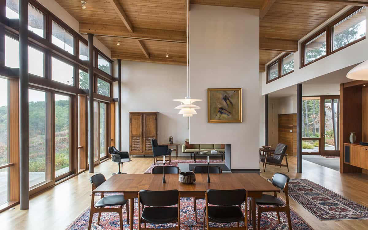 Open great room with exposed wood ceiling beams, walnut dining table with black chairs, pendant light, and floor-to-ceiling windows on two sides