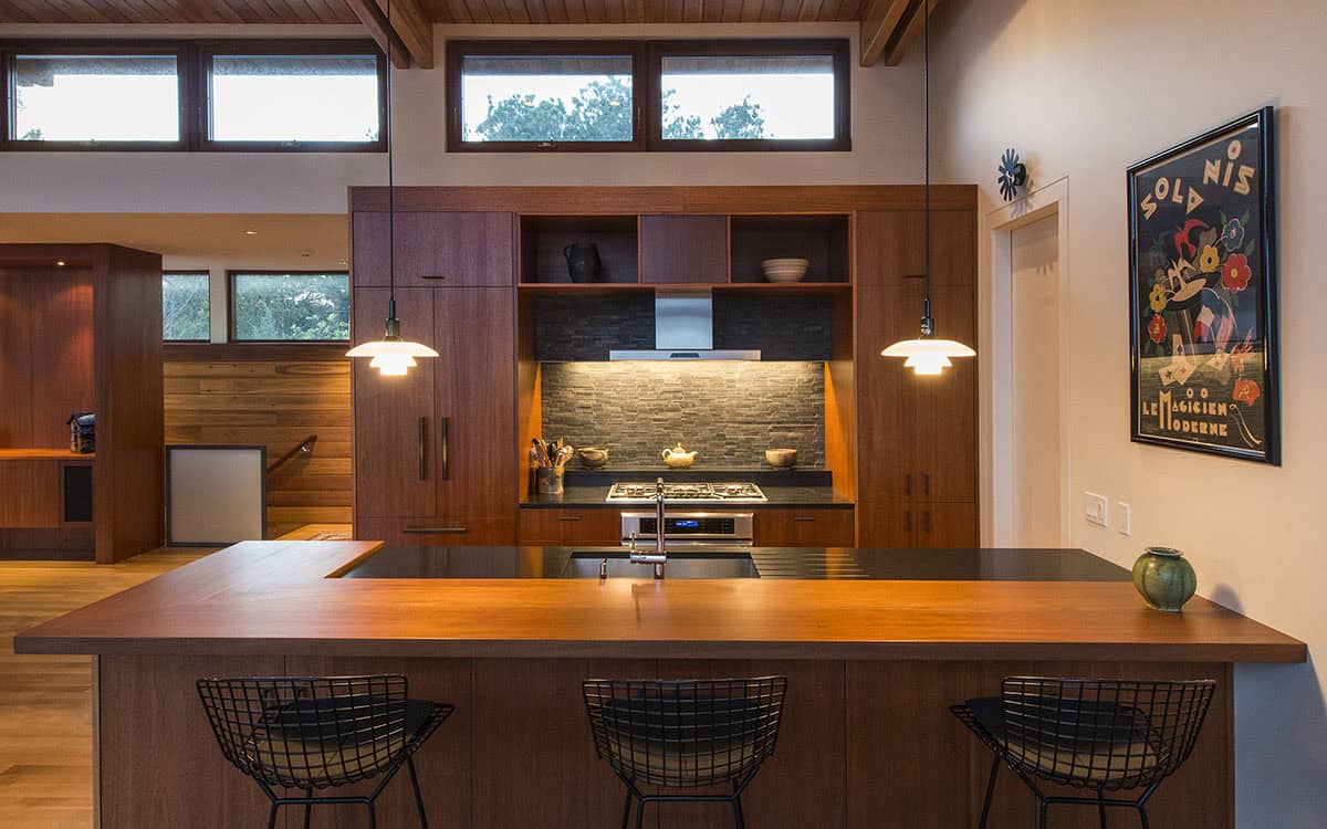 Mid-century modern kitchen with rich walnut cabinetry, stone tile backsplash, dark countertops, wire barstools, and pendant lights over an island