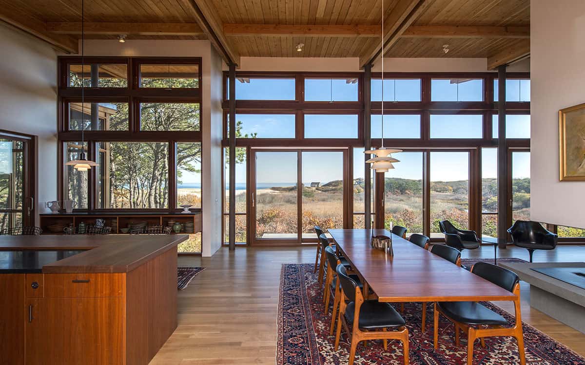 Dining area with walnut table and chairs, pendant lights, and floor-to-ceiling wood-framed windows looking out over coastal dunes and ocean